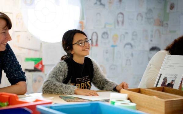 A child sits at a table with a piece of paper in front of her, smiling.