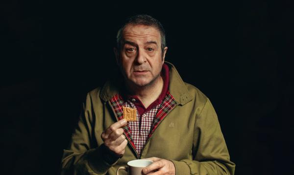 A person wearing a green jacket. They are holding a biscuit with one hand and mug in the other.