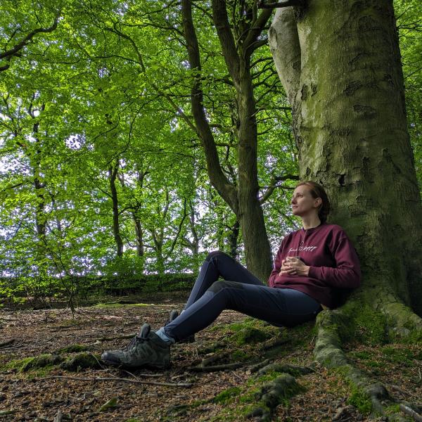 A lady sitting resting against a tree looking pensive with a mug of tea, in a green leafy woodland