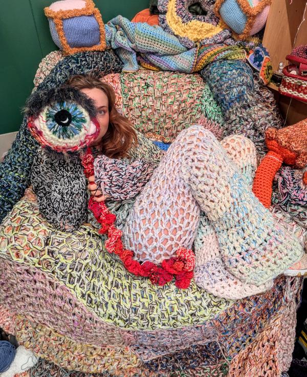 A woman sits on a crochet covered armchair, wearing a crochet costume and holding a crochet eyeball to her face