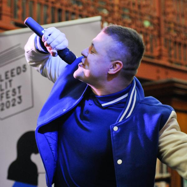 Jamie, a white male, holding a microphone up to his mouth. Wearing a blue polo top and blue and white bomber jacket