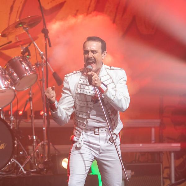 A man dressed as Freddy Mercury in a white military-style jacket singing passionately into a microphone on a stand.