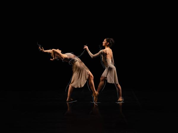 On a black background, two dancers face each other. One reaches forward with one hand while the other arches back with arms overhead.