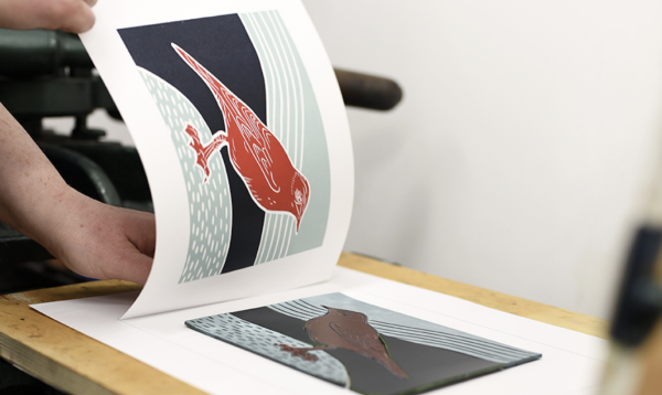 A linocut image of a bird in red ink with a blue background being revealed after printing from a pice of lino.