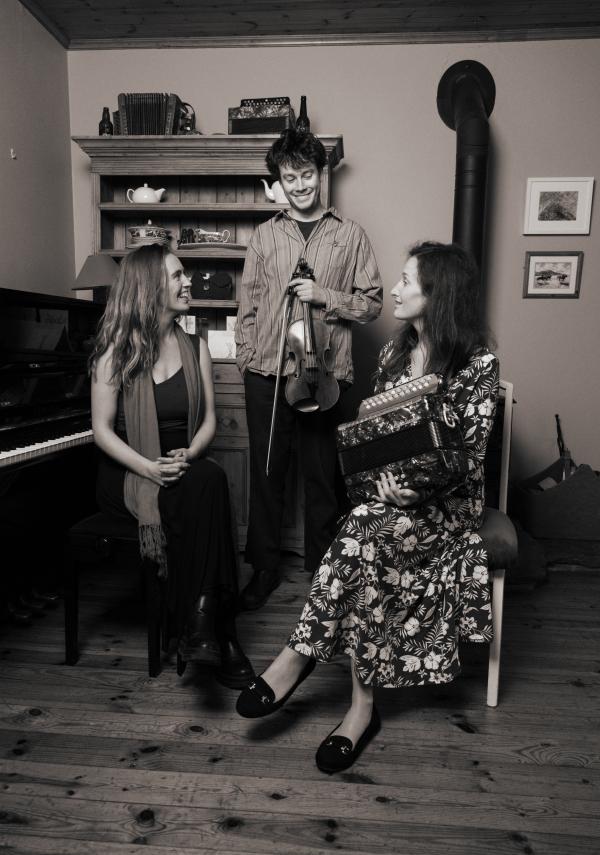 Black and white image shows a male fiddle player with a female accordian player and a female pianist.