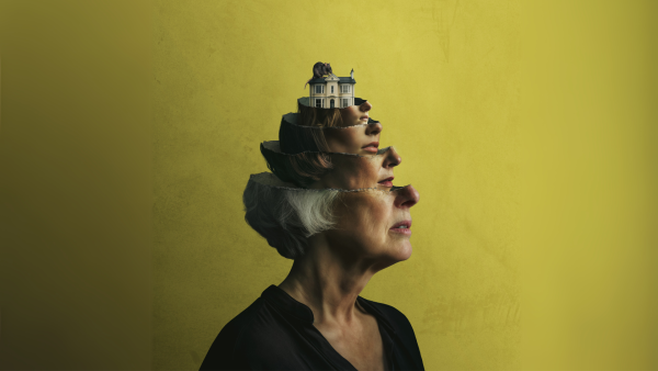 A person with grey hair and a black shirt. The head makes a matryoshka of the same face but younger and younger. At the centre, a miniature house.