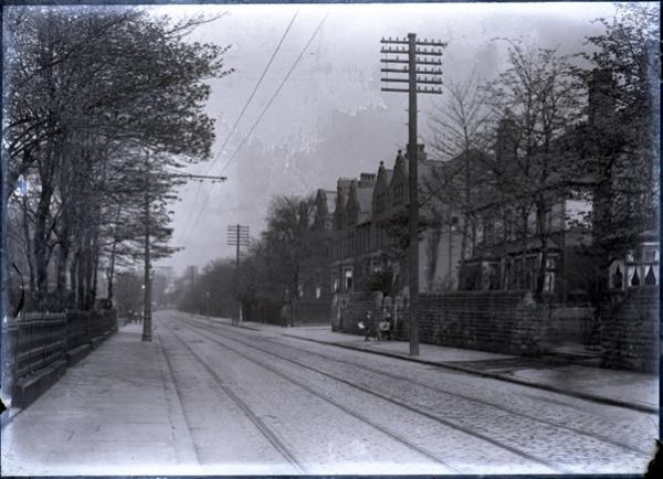 A black and white photo of a street with tramlines down the middle. Houses and trees line the road.