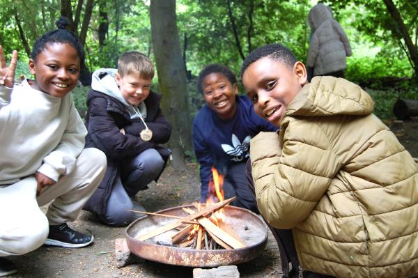 Three children in the woods, sitting round a campfire, smiling at the camera.