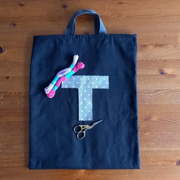 A blue tote bag which has a letter T laid on it in light blue fabric, there are two skeins of thread (light pink and dark pink) and a pair of embroidery scissors also on the tote bag.