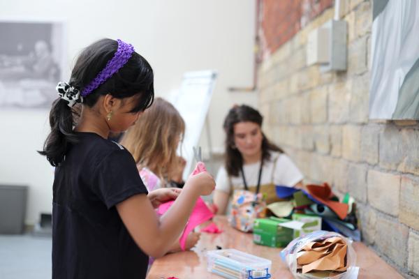Image shows young girl working on a textile piece on a table, another young girl is in the background along with workshop facilitator Sonia. 