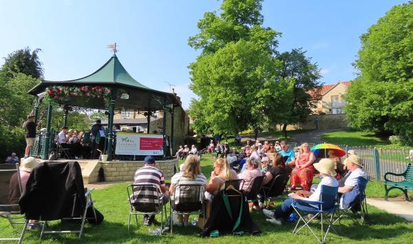 A sunny day with a crowd of people sat on deck chairs facing a band stand. There is a brass band performing on the bandstand.