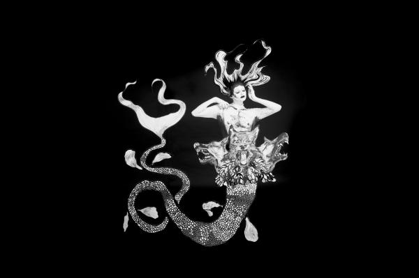 A monochromatic mermaid stands against a plain black backdrop. Her eyes are closed, hands framing her face and has long flowing hair. Her tail is long and spindly with three snarling dog heads placed at her waist.
