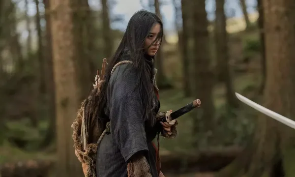 A bedraggled woman with long dark hair, and a katana by her side, stands in a forest. An unseen person holds a sword pointed towards her.