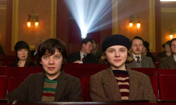 Hugo and his acolyte sit in the cinema, facing the camera. Adults sit behind them.
