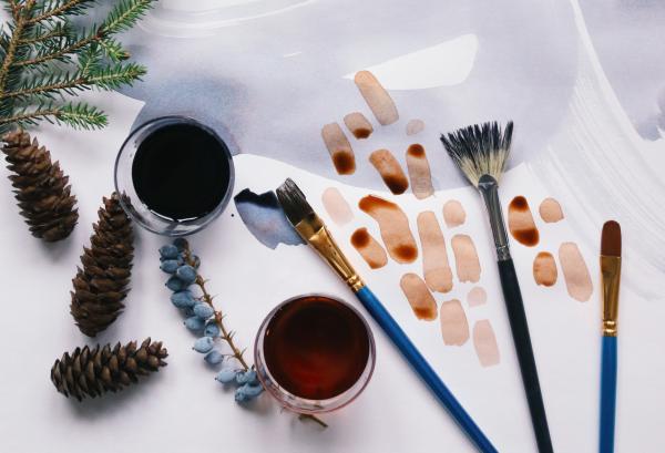Art supplies and natural elements on a white surface, including paints, brushes, pinecones, and evergreen branches.