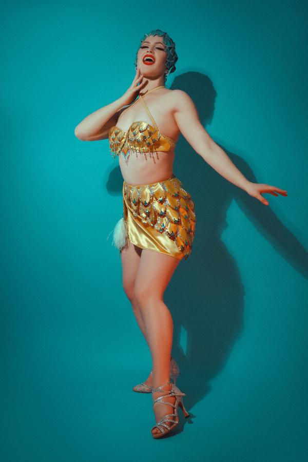 Picture of Aurora Nova, a showgirl wearing an embellished yellow costume in front of a turquoise backdrop