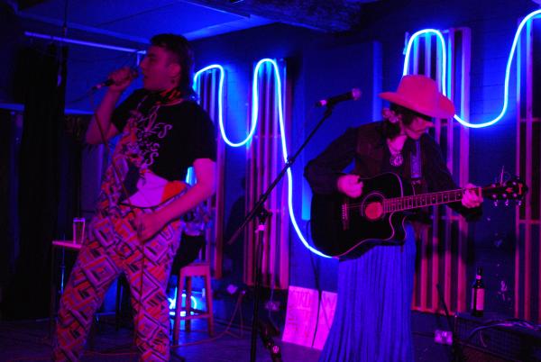 A photo of ThemTheres Cowboys performing at wharf. Lewis is in their dungarees with a colourful pattern and Spike is wearing a pinkc owboy hat, a blue skirt and carrying their guitar.