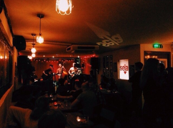 A band plays to a busy room on a red lit stage.