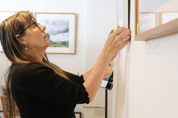 A white lady with long brown hair and glasses is adjusting a picture in a frame on the wall