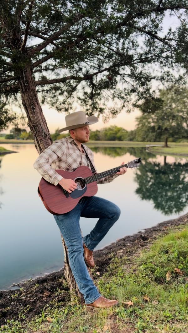 Owen Morton Holding a guitar stood by a tree and a lake.