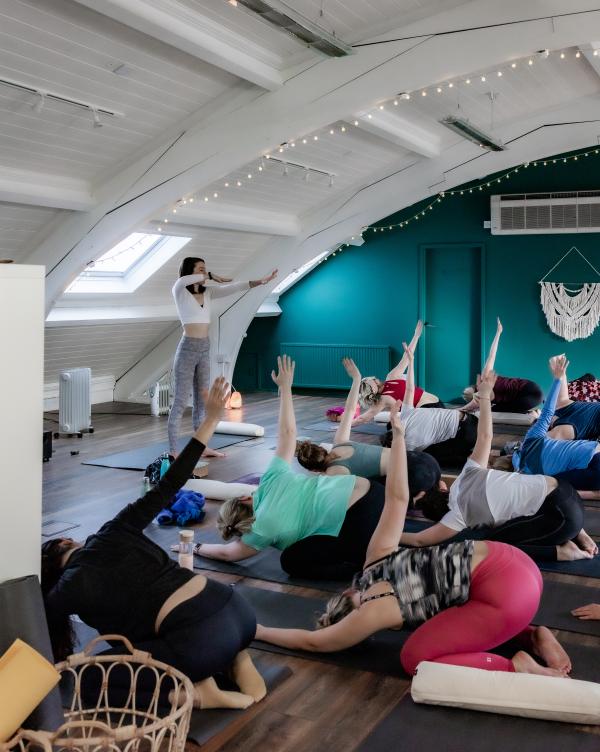 A yoga teacher guides a class in a shoulder stretch in a studio with fairy lights and arched ceiling.