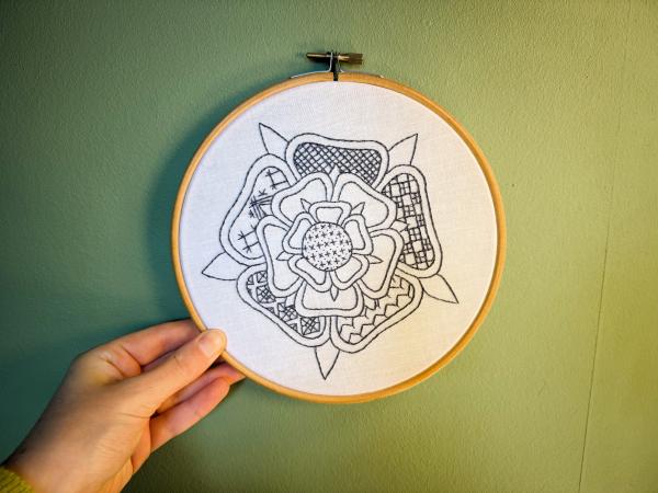 A hand holds a black embroidered rose design in an embroidery hoop against a green background.