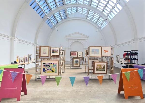 Framed pictures on stands in a large white room with a glazed ceiling, there is colourful bunting and information boards in the foreground