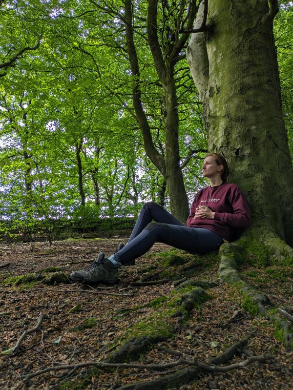 A lady sat at the base of a tree in a woodland holding a mug and looking reflectively