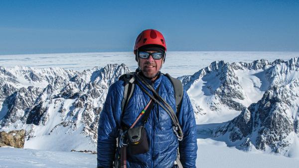 A man in climbing gear stands with a background of snow-covered mountains.