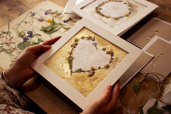 A person holds a framed artwork of dried flowers forming a heart, surrounded by other botanical art.