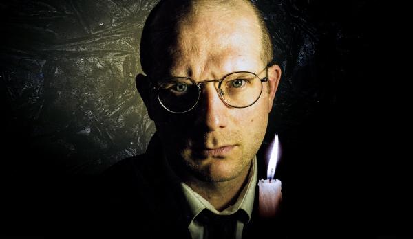 A man holding a candle in a dark room looks intensely into the camera.