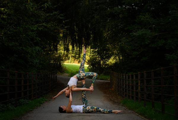 wo Acro Yoga performers in a path, in a park covered by trees. They are mid pose, one is on the ground holding the other one up