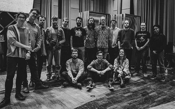 A black and white group shot of the Ancient Infinity Orchestra on a stage, featuring 15 members