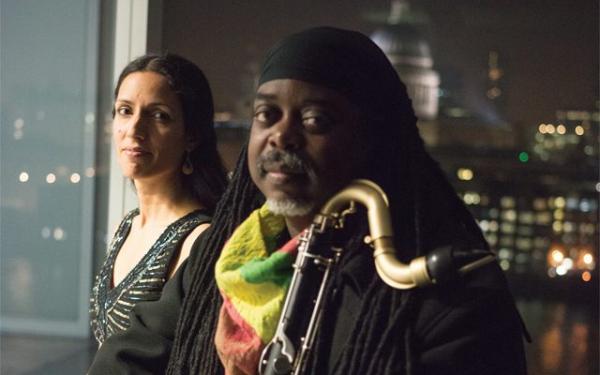 A man holding a saxophone, stood next to a woman with the US Capitol Building visible in the background through a window