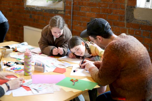 Image shows two children and man sat drawing at a table. The table is filled with scrap paper and drawing materials. 