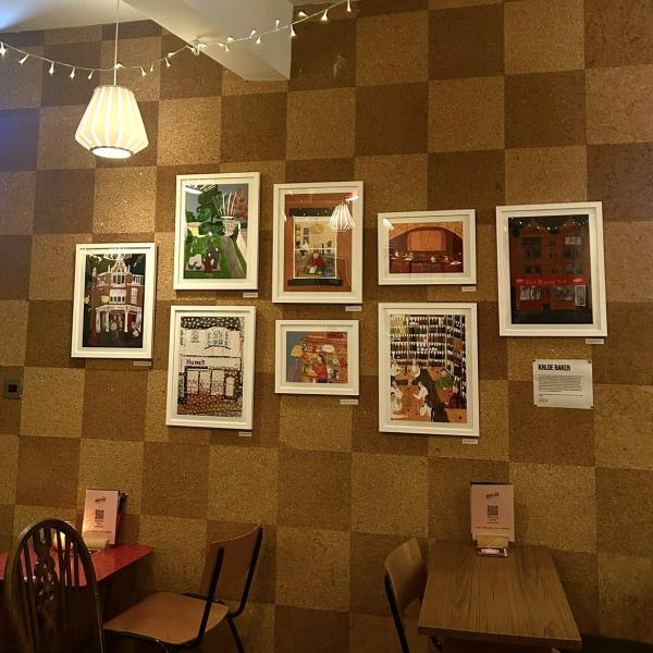 Ten gouache illustrations in white frames hung on a corkboard wall in a bar space. The room is dimly lit with fairy lights and candles. 