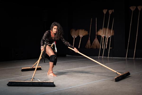 Venuri Perera, a young Sri Lankan woman dances under a spotlight. Her long brown curly hair cascades over the left side of her face as she stares into the distance. She is wearing a black lace leotard and black knee pads. Around her neck is a thick beaded necklace in red. As she hunches she pushes two brooms forward. The brooms are made out of wood and are long and wide. She has a broom tucked behind her, between her legs.