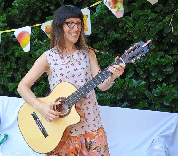 An image of a lady (Nicky Bray) with a guitar, smiling, in a garden with bunting in the background.