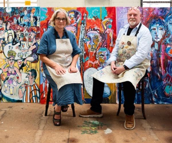 Photo shows two Artists, Mel Davies and Kim Coley, Wearing paint  stained aprons, seated in front of large scale and colourful art works.