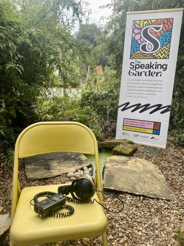 In an outside space with foliage in the background, a yellow chair has headphones and dictaphone on it. In the background is a banner for the Speaking Garden.