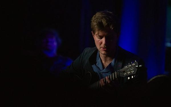 Tom Ollendorff playing guitar on a darkly lit stage