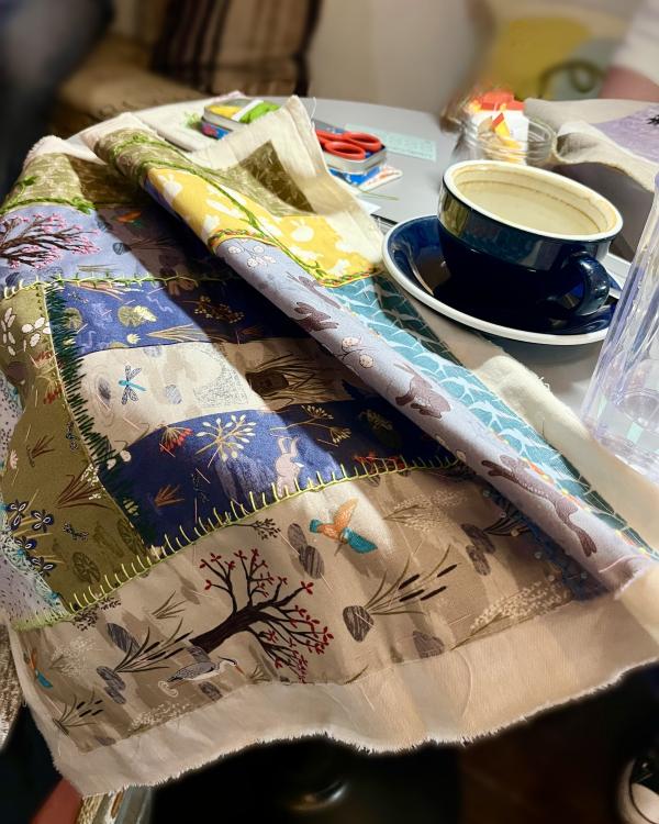 A table with a patchwork hand stitched cloth and a cup and saucer to the right