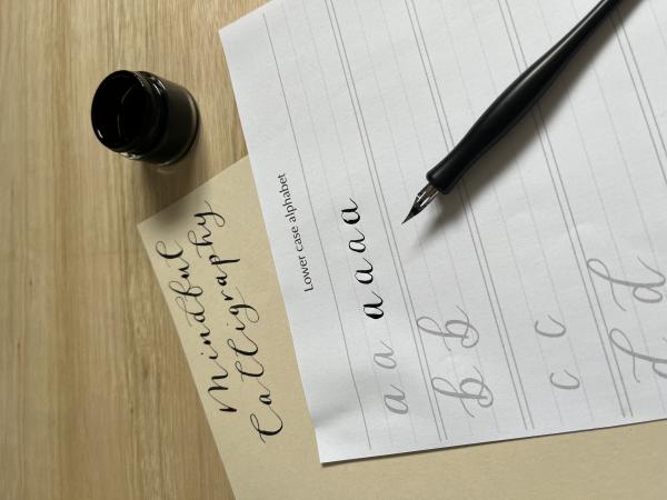 Image shows a piece of paper for practice with modern calligraphy letters. A traditional calligraphy nib and a pot of ink rest on top of the paper