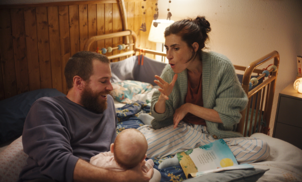 A man and a woman sit on a bed with a baby. The woman faces towards the camera and is shown speaking with sign language.