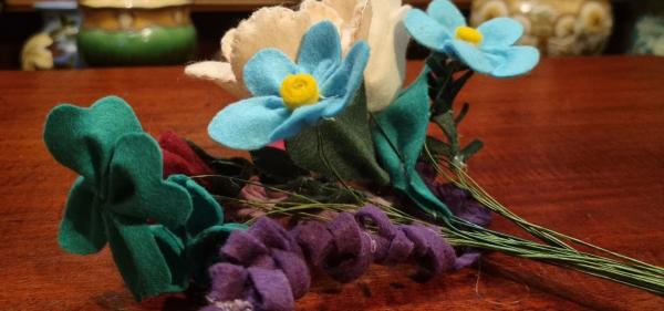 An image of some flowers made of felt. They are purple, green, white, and blue with yellow centres. They are laid on a wooden counter top.