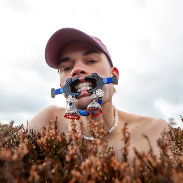 Head view of Herbie, shoulders up with a bush in front of him and under his head. He is wearing baseball cap and a device that stretches his lips to bare his teeth
