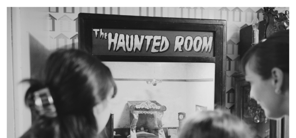 A black and white picture of a family looking at a penny arcade machine called 'The Haunted Room'.