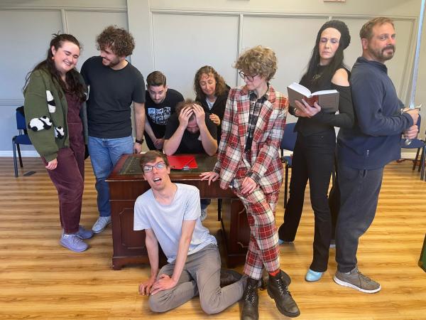 A group is gathered around a desk. To the right a woman stands reading a book. Her back is against a man holding a sword. A man and a woman stand on the opposite side of the desk close and smiling at each other. There is a man on the floor looking scared and another man with his head in his hands sat at the desk. Behind him, three other people look concerned for him.