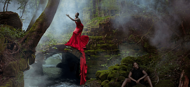 A woman in a wood wearing a red dress stood on a rock, the same woman wearing black sat on the mossy ground