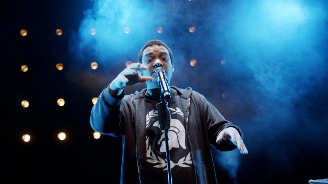 A man in dark clothes is speaking into a microphone in front of a black backdrop. Dramatic spot lights and blue smoke fill the space.